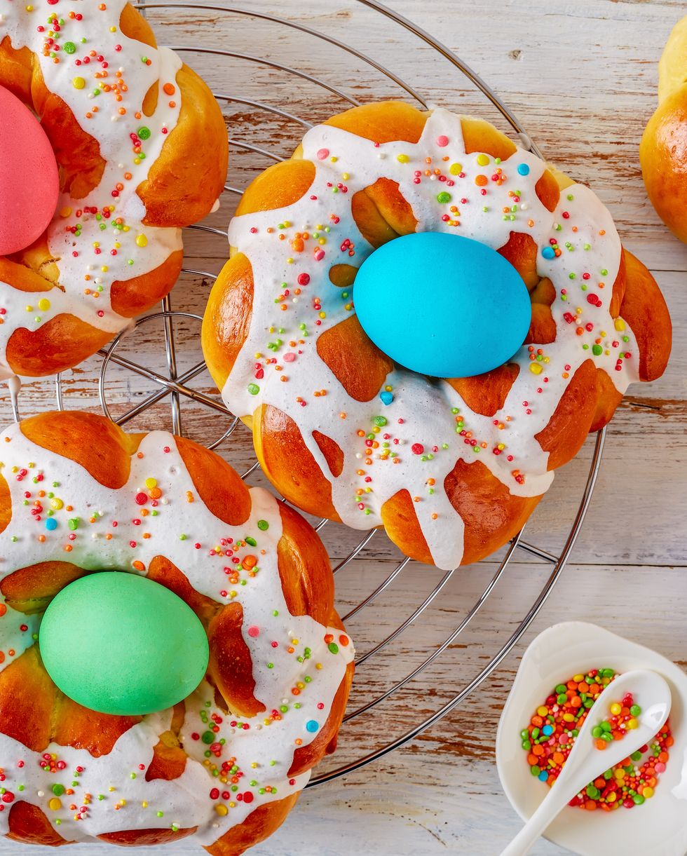homemade sweet italian easter bread rings glazed around dyed egg and topped with colorful sprinkles on a wire rack, on an old white wooden table, view from above, flatlay