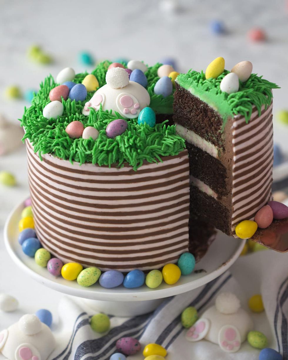 easter bunny cake with striped frosting and piped grass on top with speckled egg candy and a bunny sticking out from the grass