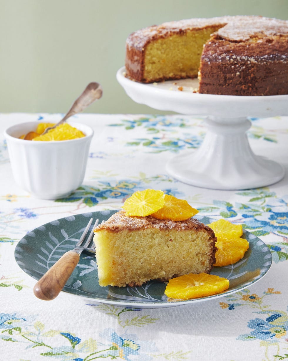 a slice of olive oil cake with citrus compote on a dark green floral plate with the rest of the cake on a white cake stand in the background