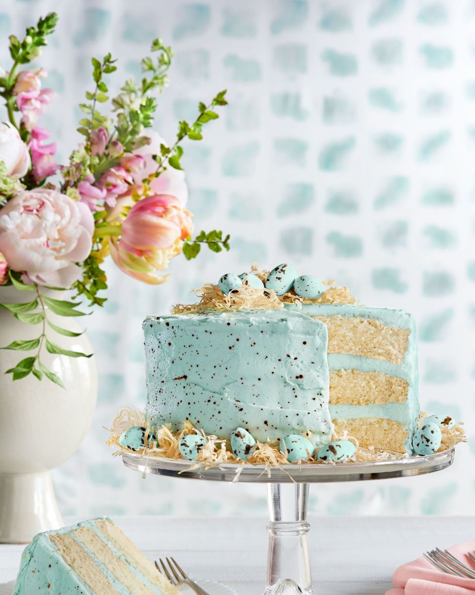 three layer malted coconut cake with light blue speckled frosting on a glass cake stand and garnished with speckled egg candies