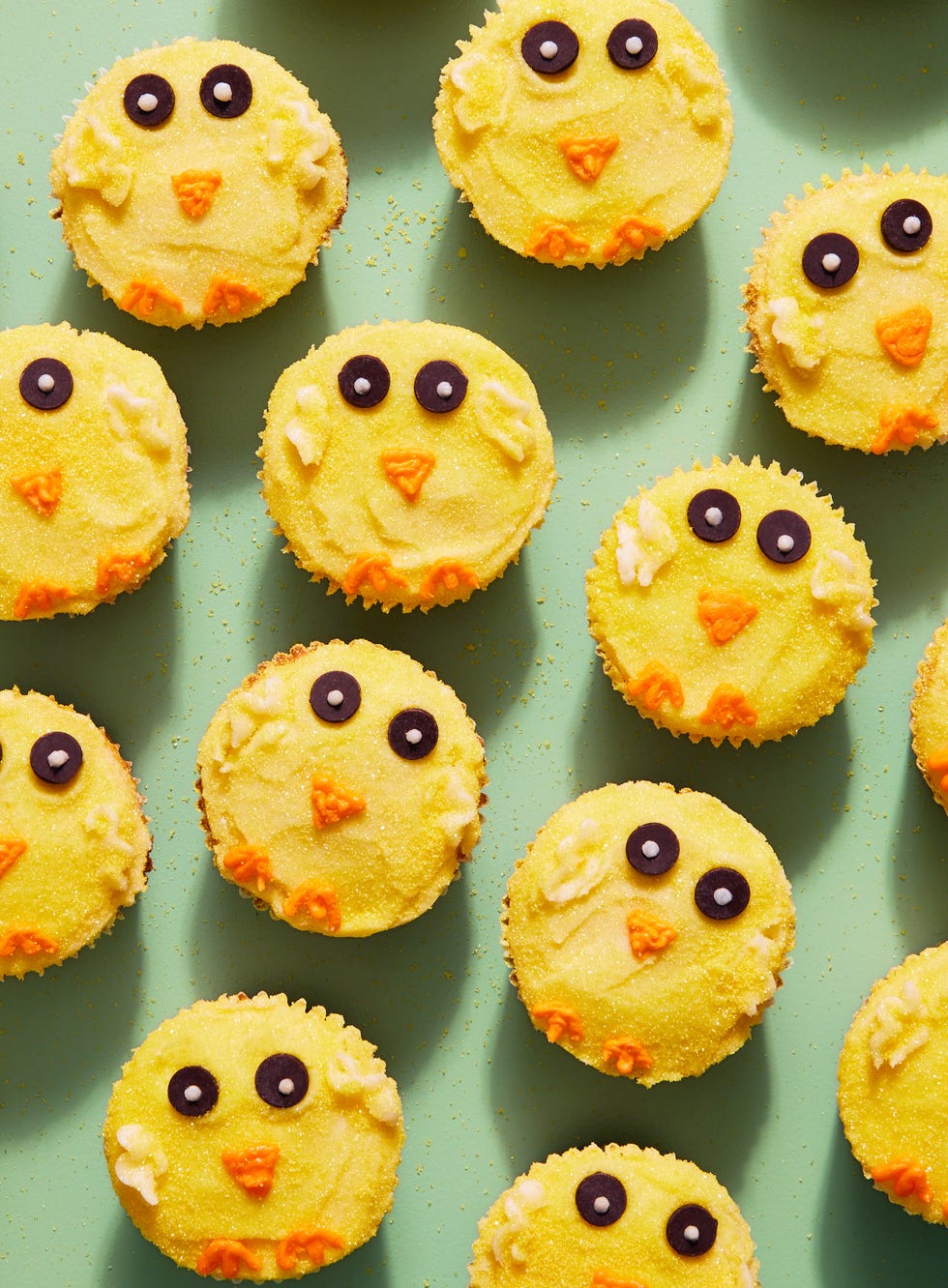 cupcakes with yellow frosting and sanding sugar decorated to look like easter chicks with chocolate chip eyes and an orange frosting beak and feet