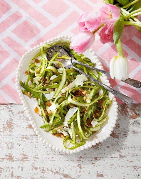 shaved asparagus and parmesan salad with raisins in a white serving bowl with serving utensils propped on the side