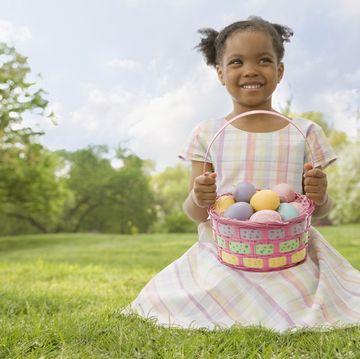 little girl smiling in green field wearing pastel plaid dress with basket full of colorful eggs found during easter egg hunt