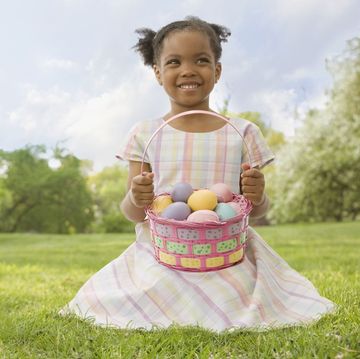 little girl smiling in green field wearing pastel plaid dress with basket full of colorful eggs found during easter egg hunt