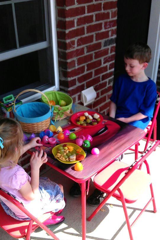 two children sitting at a red table eating lunch and playing with easter eggs