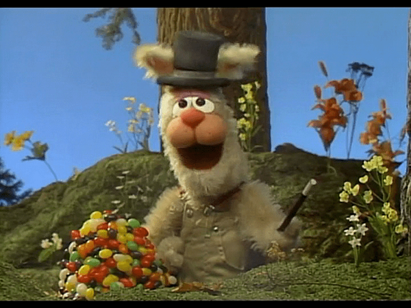 a bunny puppet wears a hat and sits in front of a giant pile of jellybeans in a scene from an easter story , a good housekeeping pick for best easter movies for kids