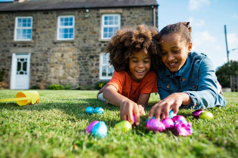 two siblings lying down in the grass on their front smiling while holding their easter eggs