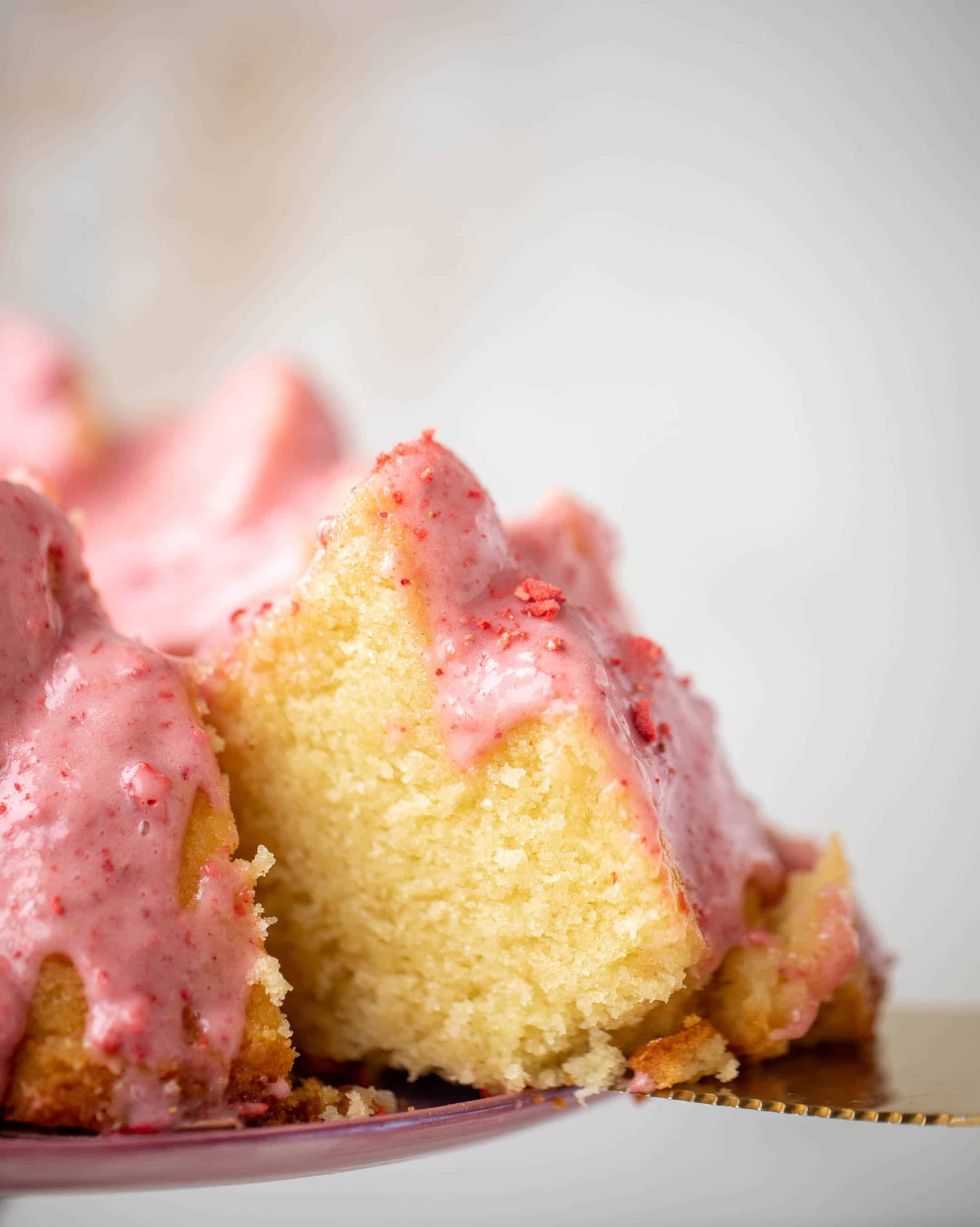 pound cake on a cake stand with strawberry icing and a cake lifter removing a slice
