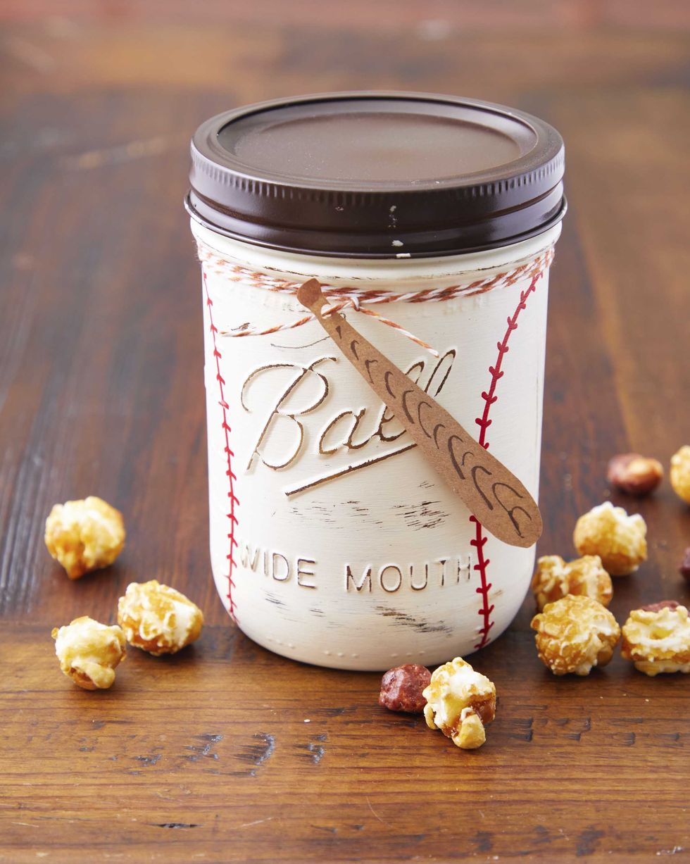 a mason jar decorated to look like a baseball with a tag made to look like a bat set up a wood table with popcorn scattered around it