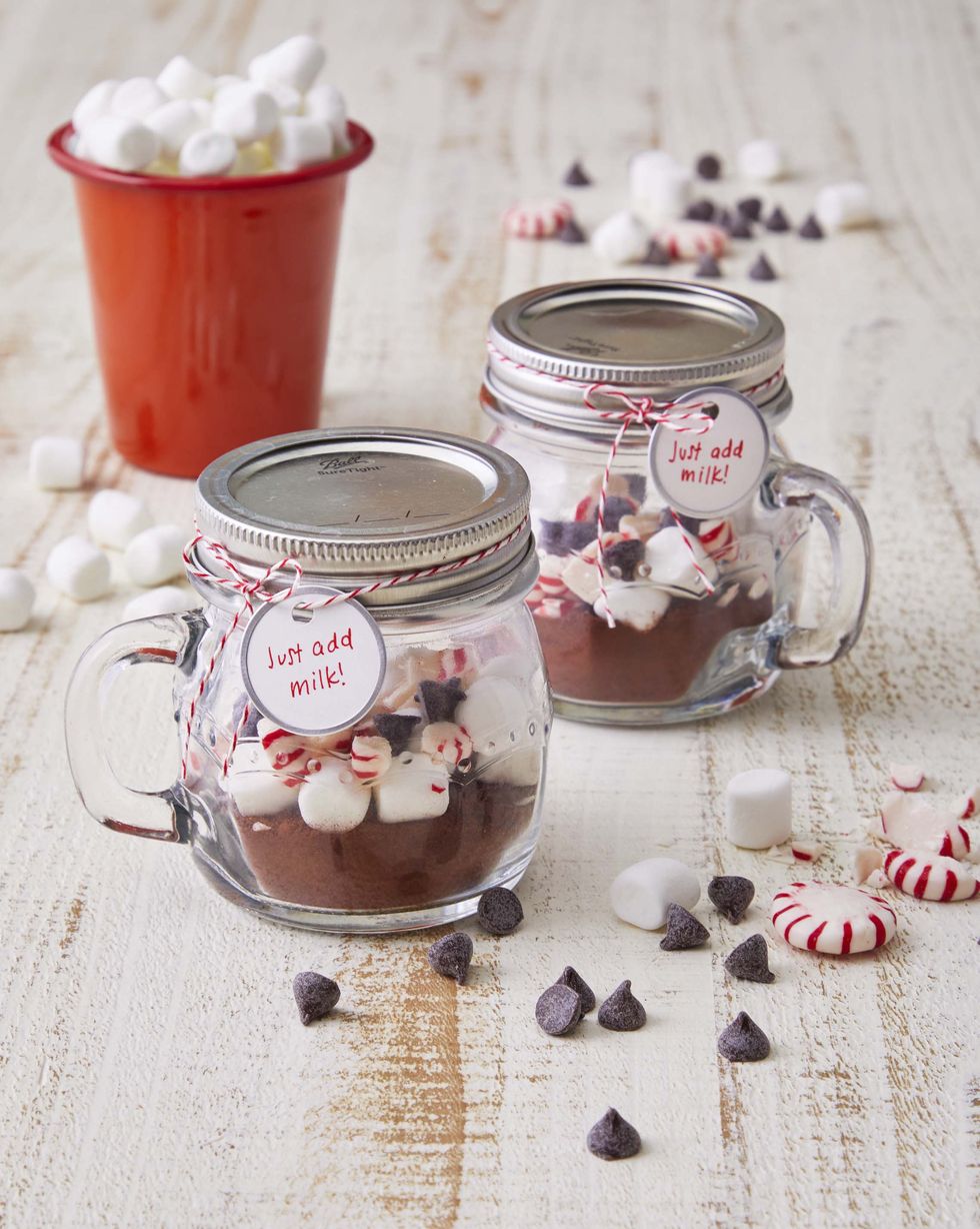 a small mug style mason jar filled with hot cocoa and topped with marshmallows chocolate chips and crushed peppermint candies set on a white table