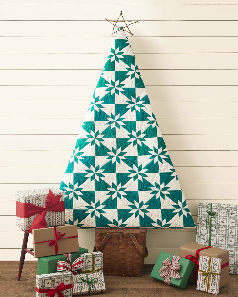 a quilt shaped into a triangle meant to resemble a christmas tree set on a wood picnic basket all surround by wrapped presentsby