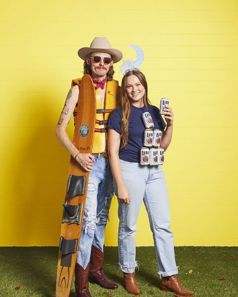 man in ripped jeans, life jacket, cowboy hat, boots holding ski, woman in tee with can pyramid glued to front, moon headband