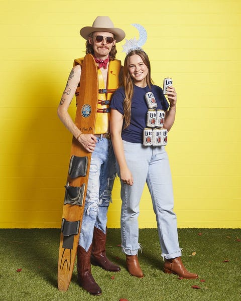 man in ripped jeans, life jacket, cowboy hat, boots holding ski, woman in tee with can pyramid glued to front, moon headband