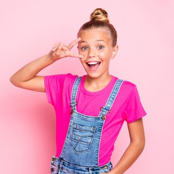 cute cheerful girl showing a peace sign in denim overalls and a pink shirt