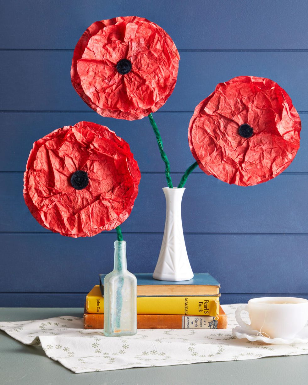 Red poppies made from coffee filters are set in a small vase in front of a blue background