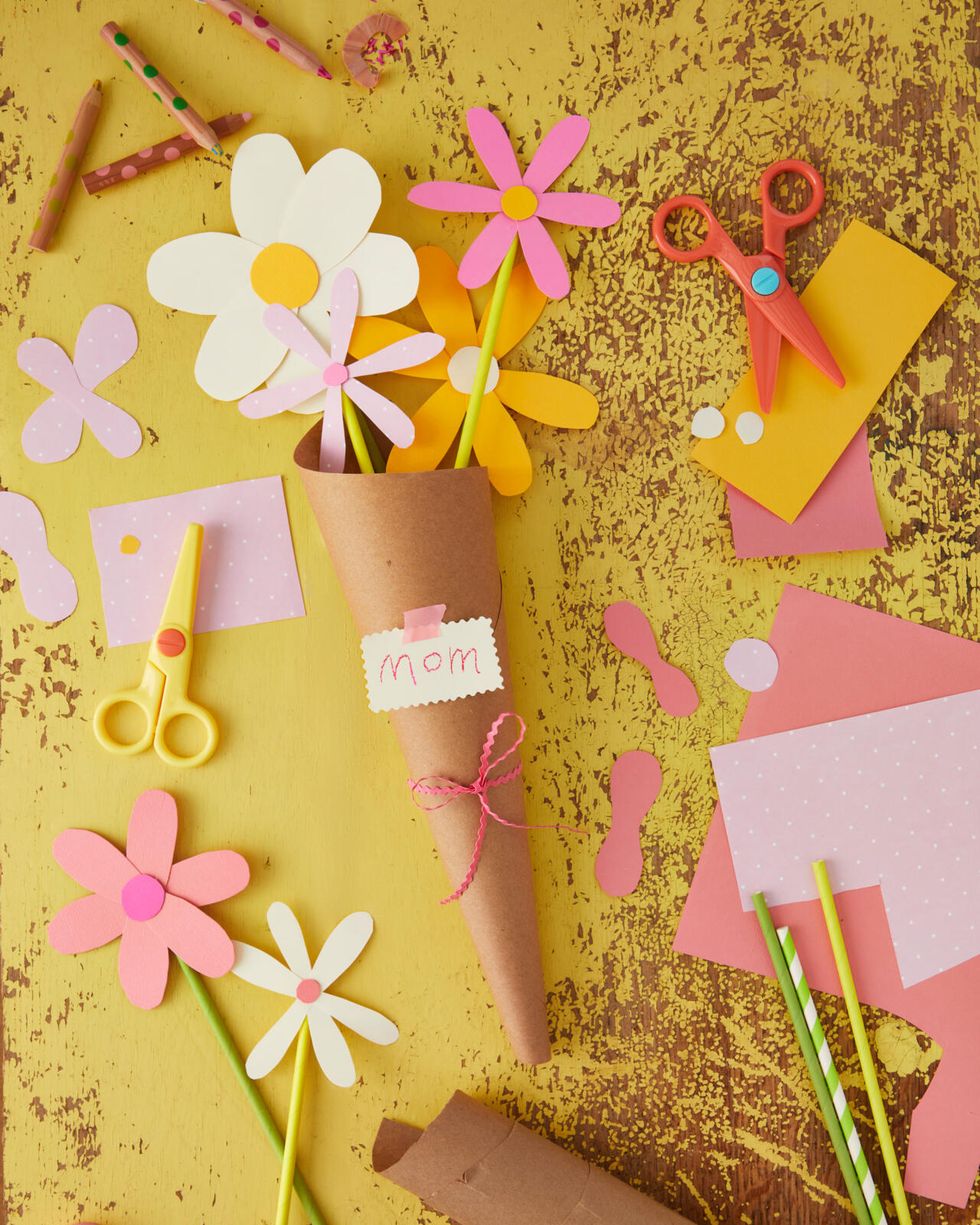 Paper flowers made of bright pastels on a yellow background arranged in a brown paper cone with a tag that says mom