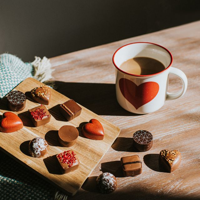 luxury chocolates in various shapes and flavours, including red heart chocolates they sit on wooden surfaces, beside a luxurious wool blanket beside the sweets sits a hot mug of coffee with a large heart emblem