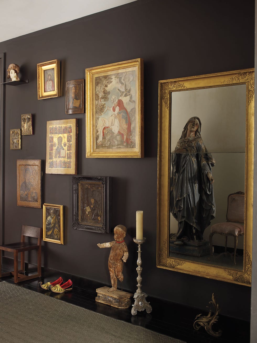 dark painted entry hall detail of a wall with multiple framed pieces of art mostly old or medieval or religious looking and some arabic looking red lined gold and black slippers on the floor next to other objects like a cherub and candlestick and chair