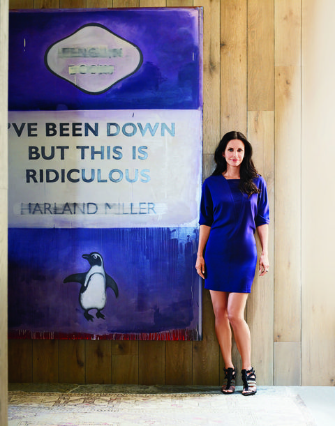 courtney cox in a purple blue dress standing next to a large book cover in the same color with the title i've been down but this is ridiculous by harland miller