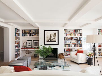 open living room painted in white with open shelves and fireplace and ottomans and glass table in the center and black and white prints