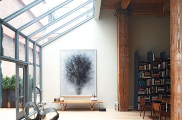 conservatory like area with wood bench with object on it and a glass wall and slanted roof adjoinging rough wood beams with furniture and loaded bookshelves