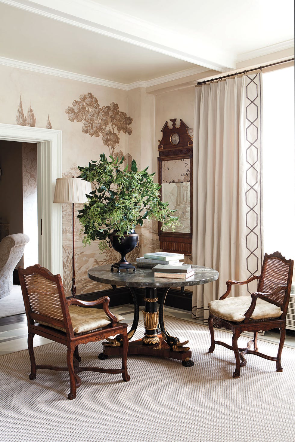cane armchairs with silk cushions around a marble top table with an antique wood base and standing lamp with white shade at the back and and urn vase with large sprigs atop the table