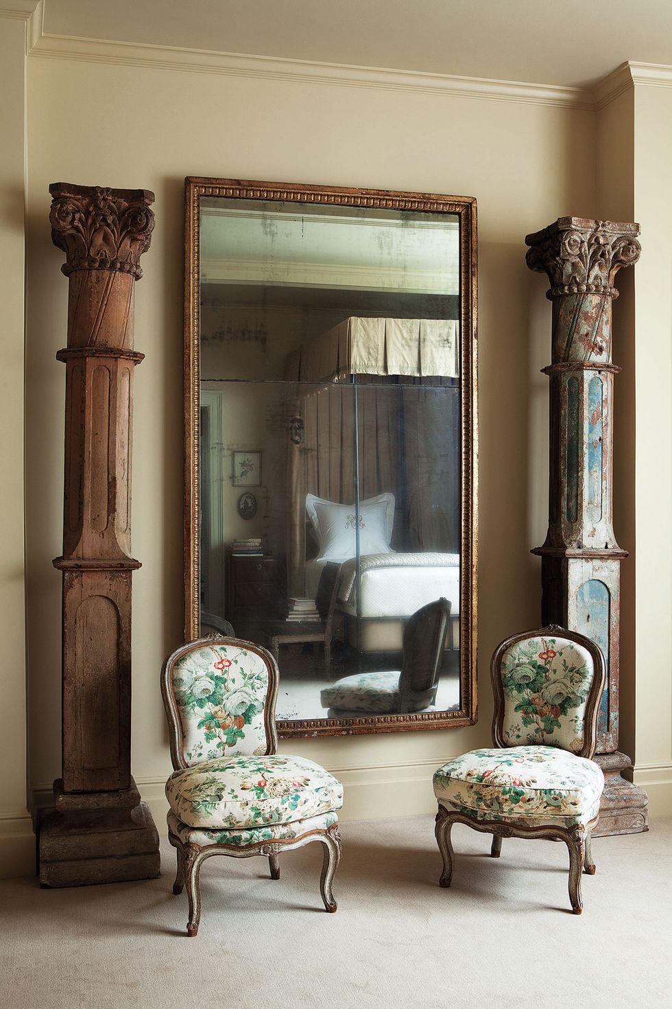two armless chairs in a floral pattern and a between in s large wood framed mirror and two tall wooden distressed columns flanking it