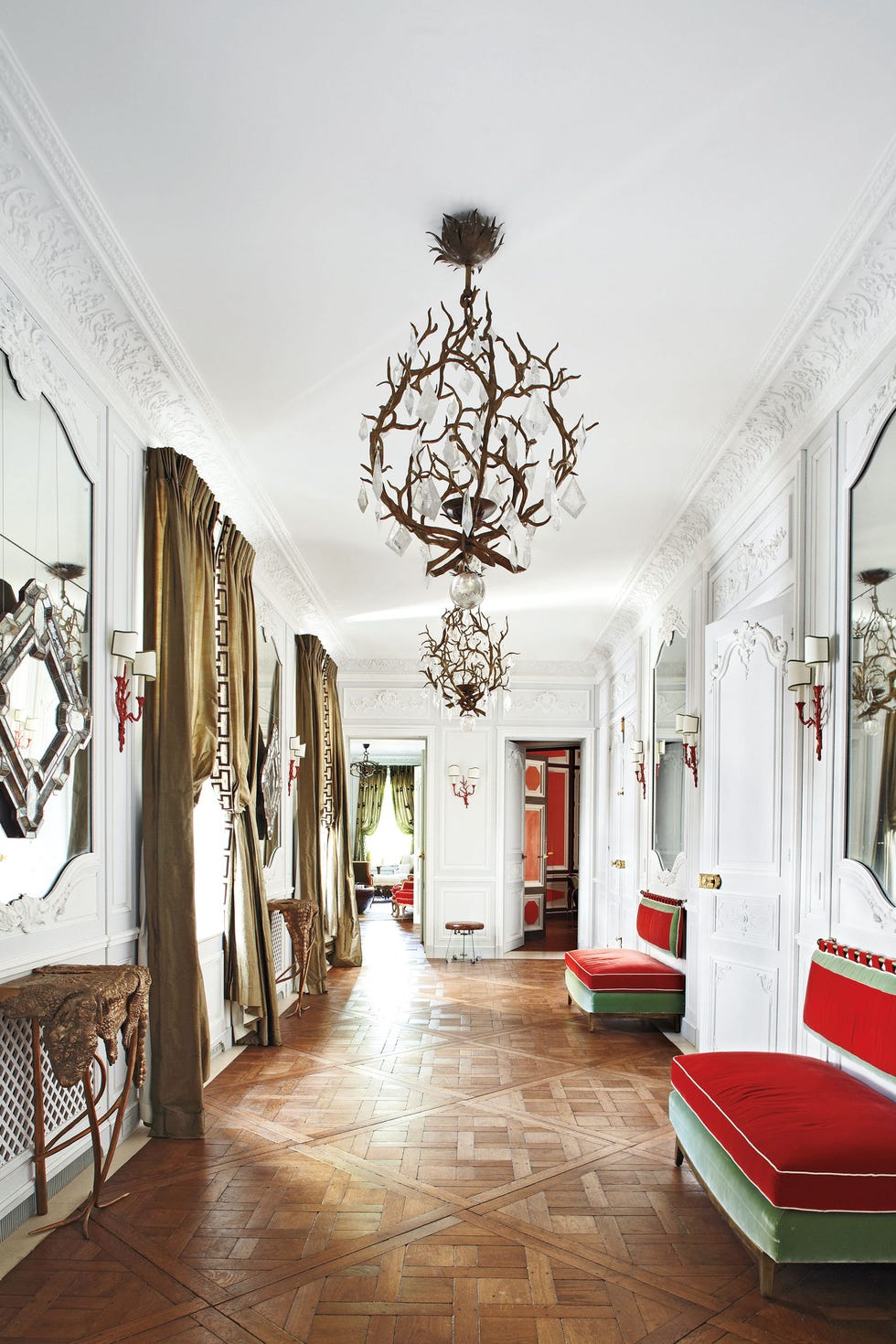 large entrance with parquet floor and red and light green velvet sofas on one side and high draped windows and small half-moon tables opposite with mirrors on the walls and a chandelier made of deer antlers in the middle