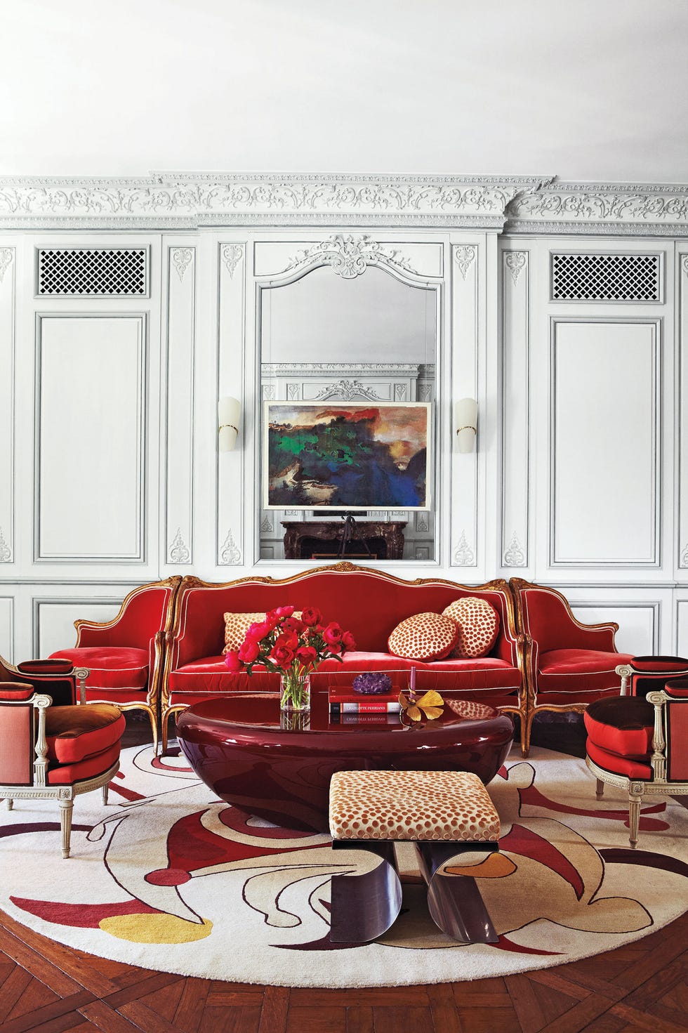 a living room with high ceilings and fancy moldings in white and a red velvet sofa with attached seats at the end and a swirling circular rug with white and red outlines and a modern leopard print stool and a shiny red lacquered bean-shaped table in the middle and a painting on the wall