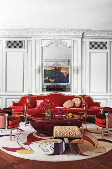 a living room with high ceilings and fancy moldings in white and a red velvet scroll sofa with end seats attached and a swirly circular white and red outline carpet and a leopard scroll leg modern stool and a shiny red lacquer bean shape table at center and a painting on the wall