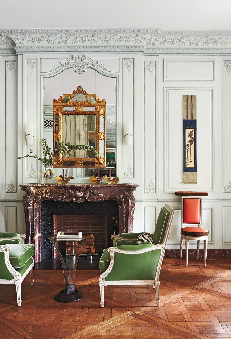 high ceilinged room with plaster molding and parquet floors and a speckled marble fireplace mantel w ith a large gold mirror set into a larger mirror and in front of the fireplace ar two green velvet upholstered and white wood chairs facing each other and a metal and glass v shaped table beween them and an orange armless chair against the wall like a soldier
