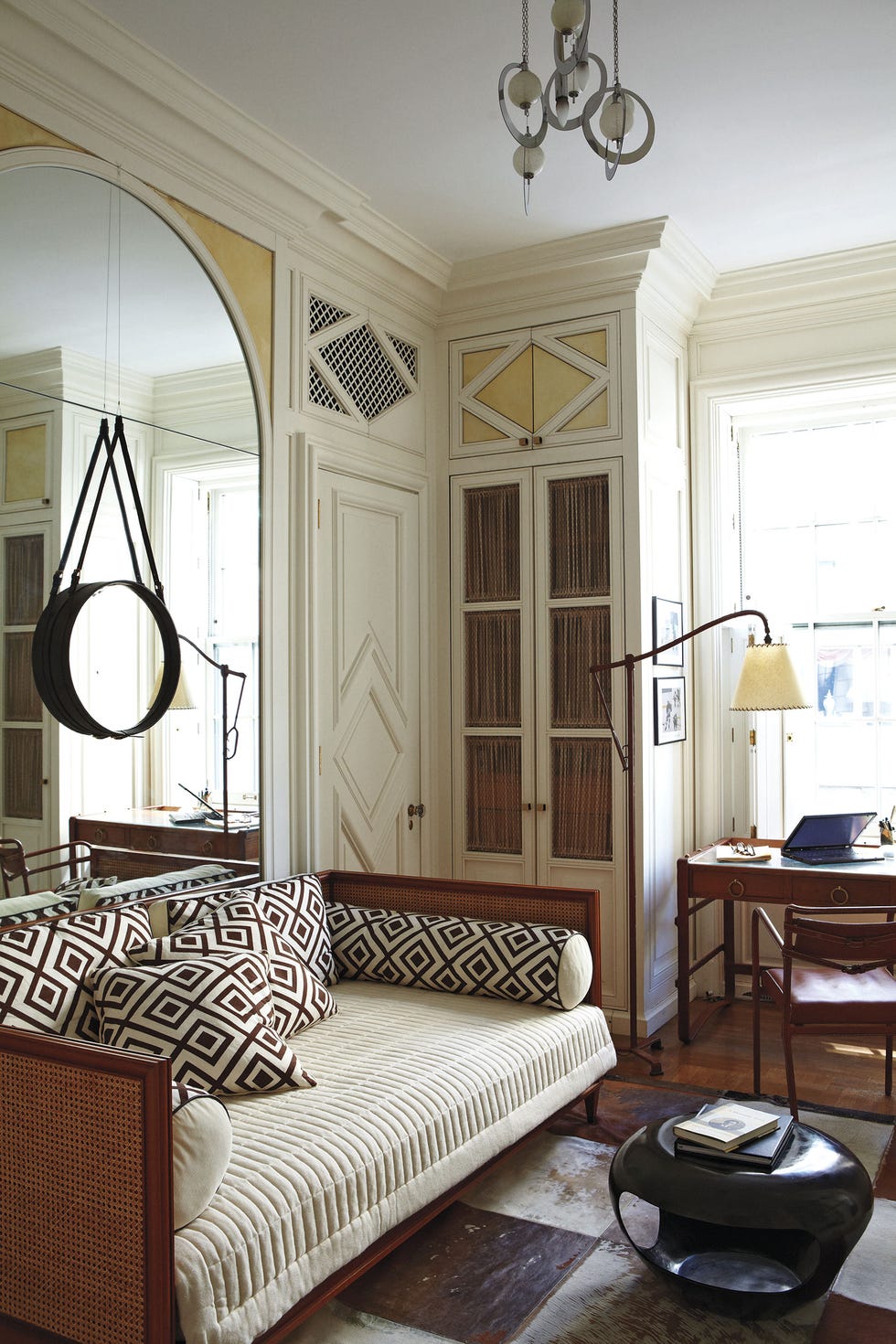 a cozy room with a rattan and wooden daybed with geometric pillows and bolsters on a light blanket and a large mirror behind it on the wall and a small black round cocktail table and a small closet in the background and a window with a desk in front of it