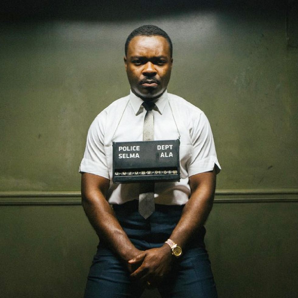 david oyelowo dressed as martin luther king jr for selma, he wears a white short sleeved collared t shirt with a silver tie and stands for a mugshot with a police board hanging around his neck