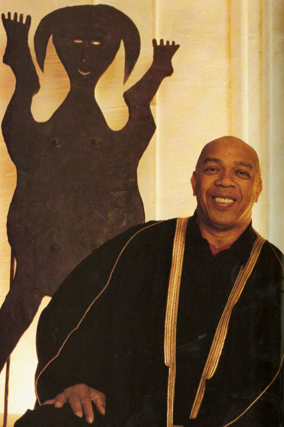 portrait of geoffrey holder in a black draped outfit with gold accents sitting in front of a large piece of metal sculpture