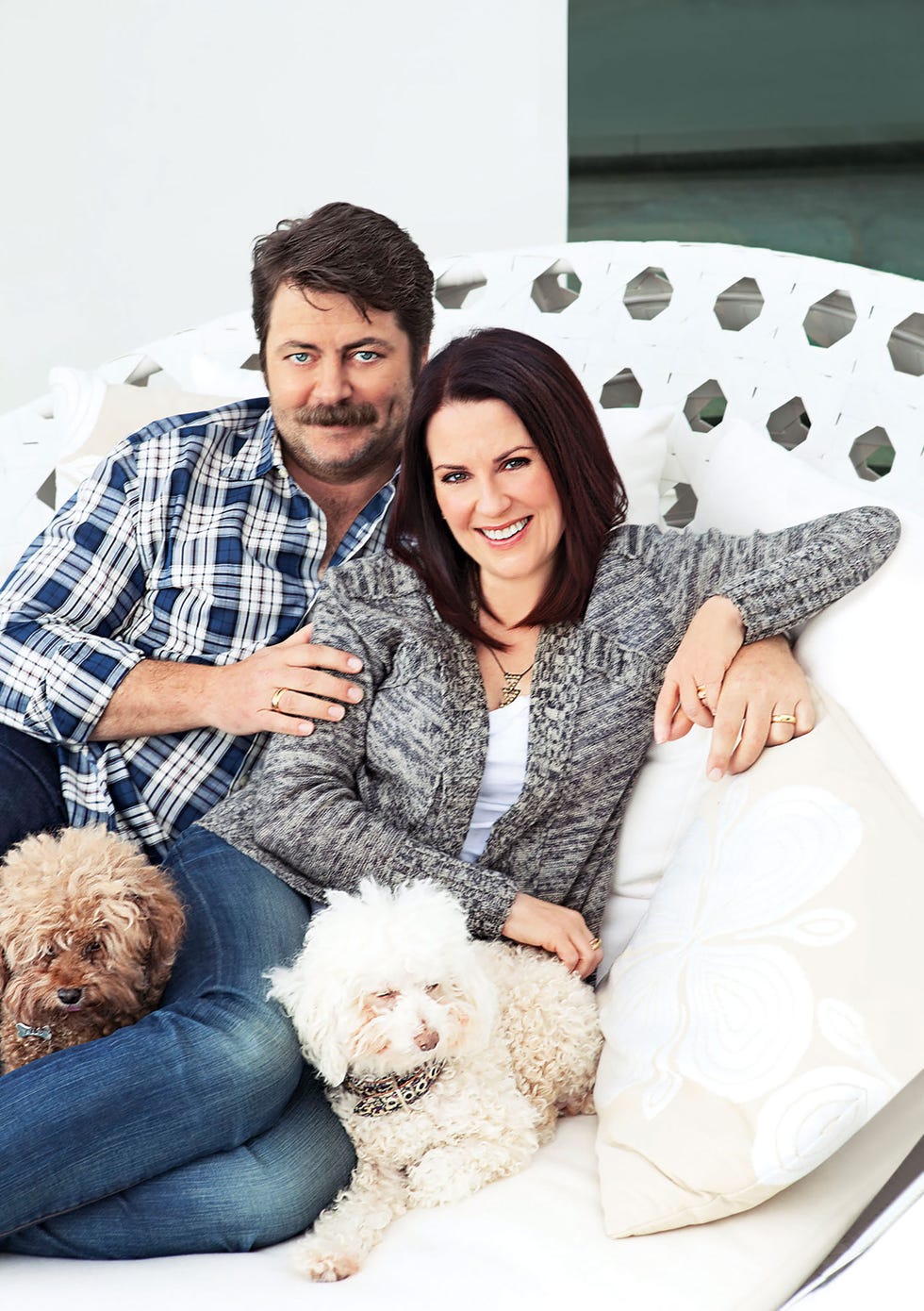 couple seated smiling into the camera and he is dressed in a plaid short and has dark brown hair and bushy mustache and piercing blue eyes and she has long straight brown hair and is wearing a comfy speckled sweater and jeans and they have their cute little dogs on their laps