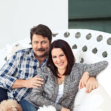 couple seated smiling into the camera and he is dressed in a plaid short and has dark brown hair and bushy mustache and piercing blue eyes and she has long straight brown hair and is wearing a comfy speckled sweater and jeans and they have their cute little dogs on their laps