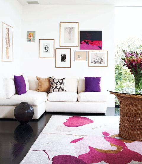 corner of a white painted room with dark floors where there is a white sofa against a wall of small framed art pieces and a wicker pedestal glass table with a floral arrangement and the rug underneath is a pale pink background ·lid with a very brilliant modernism.  the pink tulips as a design and the sofa cushions are deep purple and patterned