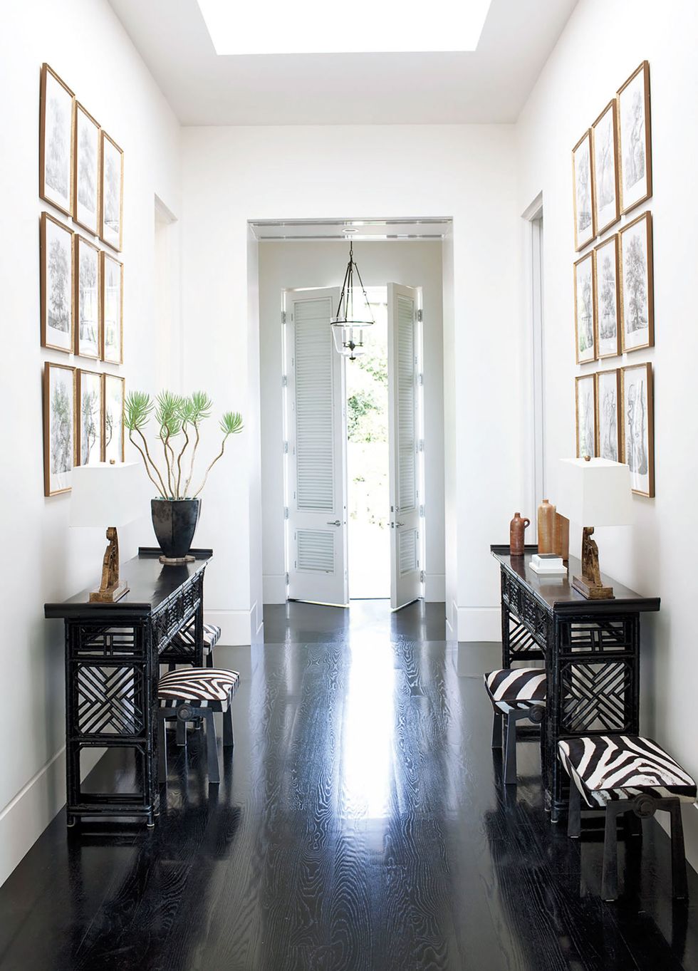 long entry way with dark floors and white walls and along the sides are thin black asian style consoles with zebra prints on the stools framed prints above them and slatted doors at the end