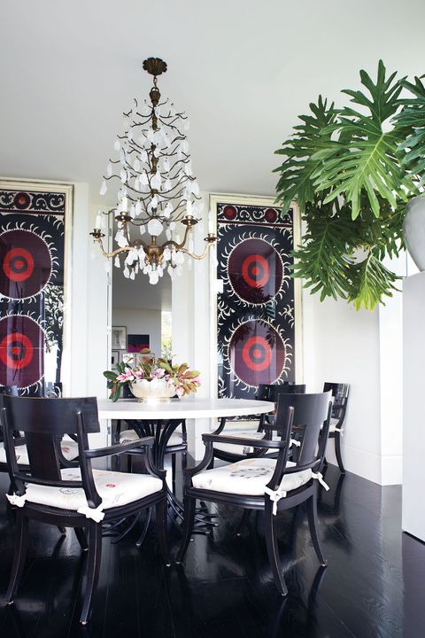 White walled dining room with dark stained floor and dark chairs paired with a twisted open black pedestal table with a white top and on the back wall are two long wide colored suzani with circular patterns.