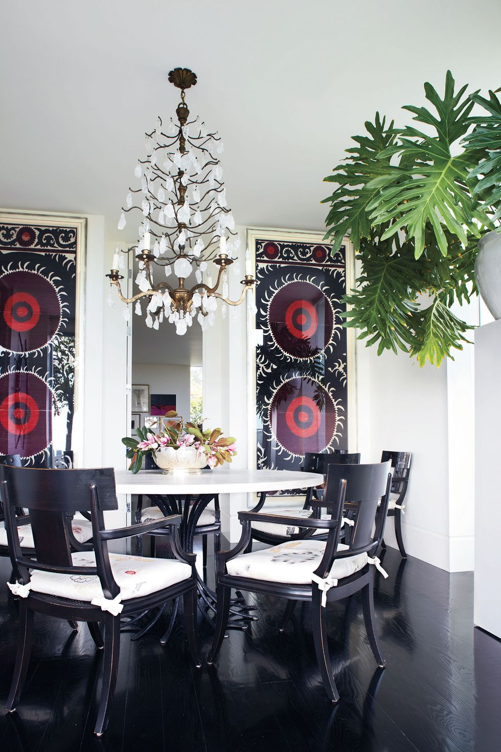 white walled dining room with dark stained floor and a dark chairs pulled up to a twisted open black pedestal table with white top and on the back wall are two long wide colorful suzanis with circular patterns