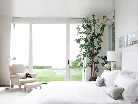 All white bedroom with a bed with white sheets with a chair by large sliding glass doors with a view outside that offsets all the white