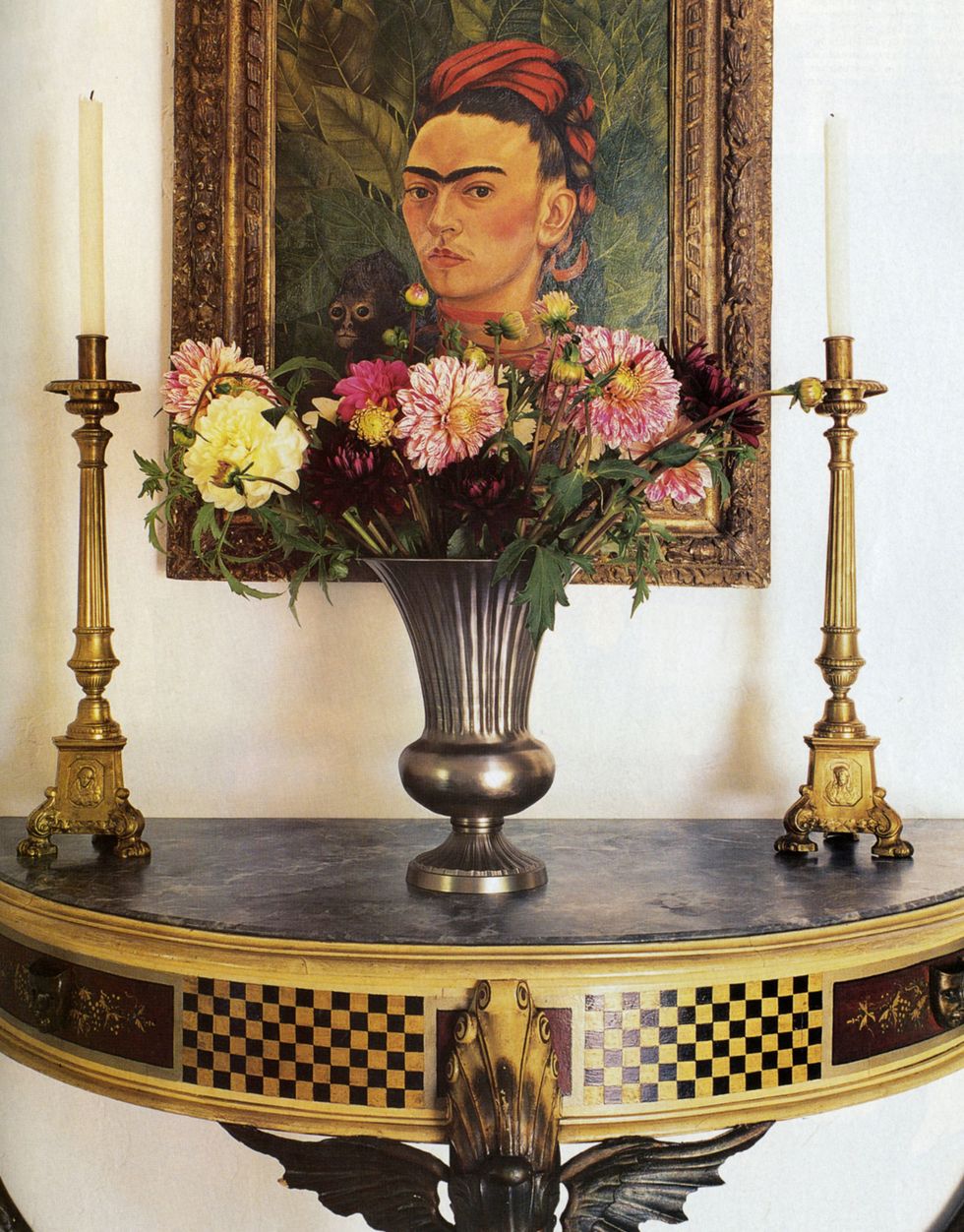 entryway with frida kahlo portrait hangs over a russian inlay demilune table with a pair of gold candlesticks and a bouquet of flowers between them