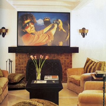media room detail of a dark terracotta tiled fireplace with black top and grate and a large painting above it of a man with sunglasses wrestling with some sort of weird demon and there is a black table at center and comfy deep cushioned chairs in a honey color with leopard spot pillows as accent on a honey yellow carpeted floor