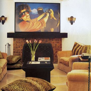 media room detail of a dark terracotta tiled fireplace with black top and grate and a large painting above it of a man with sunglasses wrestling with some sort of weird demon and there is a black table at center and comfy deep cushioned chairs in a honey color with leopard spot pillows as accent on a honey yellow carpeted floor