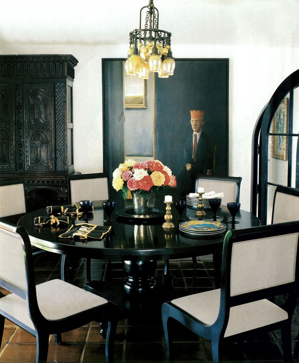 dining room with black table and black framed chair with off white seats and backs and a black armoire in the background and a dark painting featuring a skiny man in a suit wearing a red crown