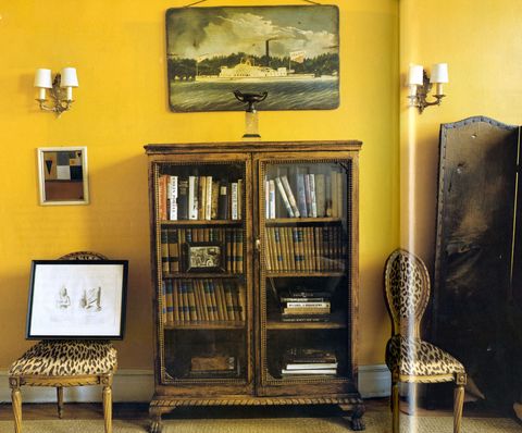room with a cheerful yellow painted wall with a old wood glass doored cabinet with books and two chairs with leopard print on either side and sconces on the wall and a screen seen on the right