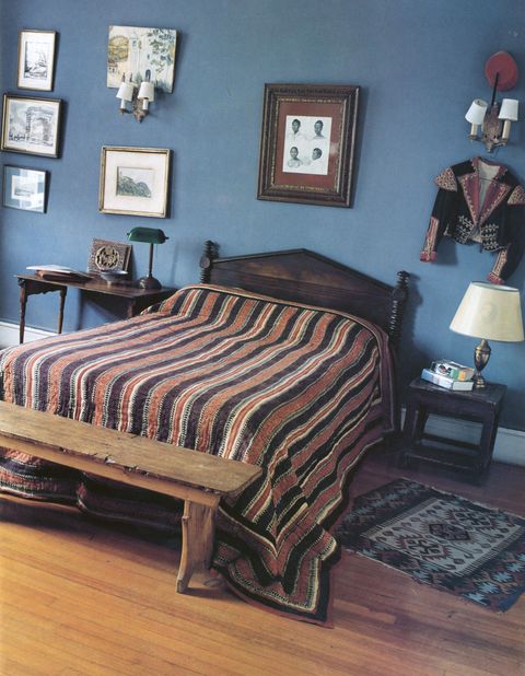 blue painted bedroom with objects hanging from the wall including a studded embroidered matador jacket and some frames and a rustic bench and striped bedcover
