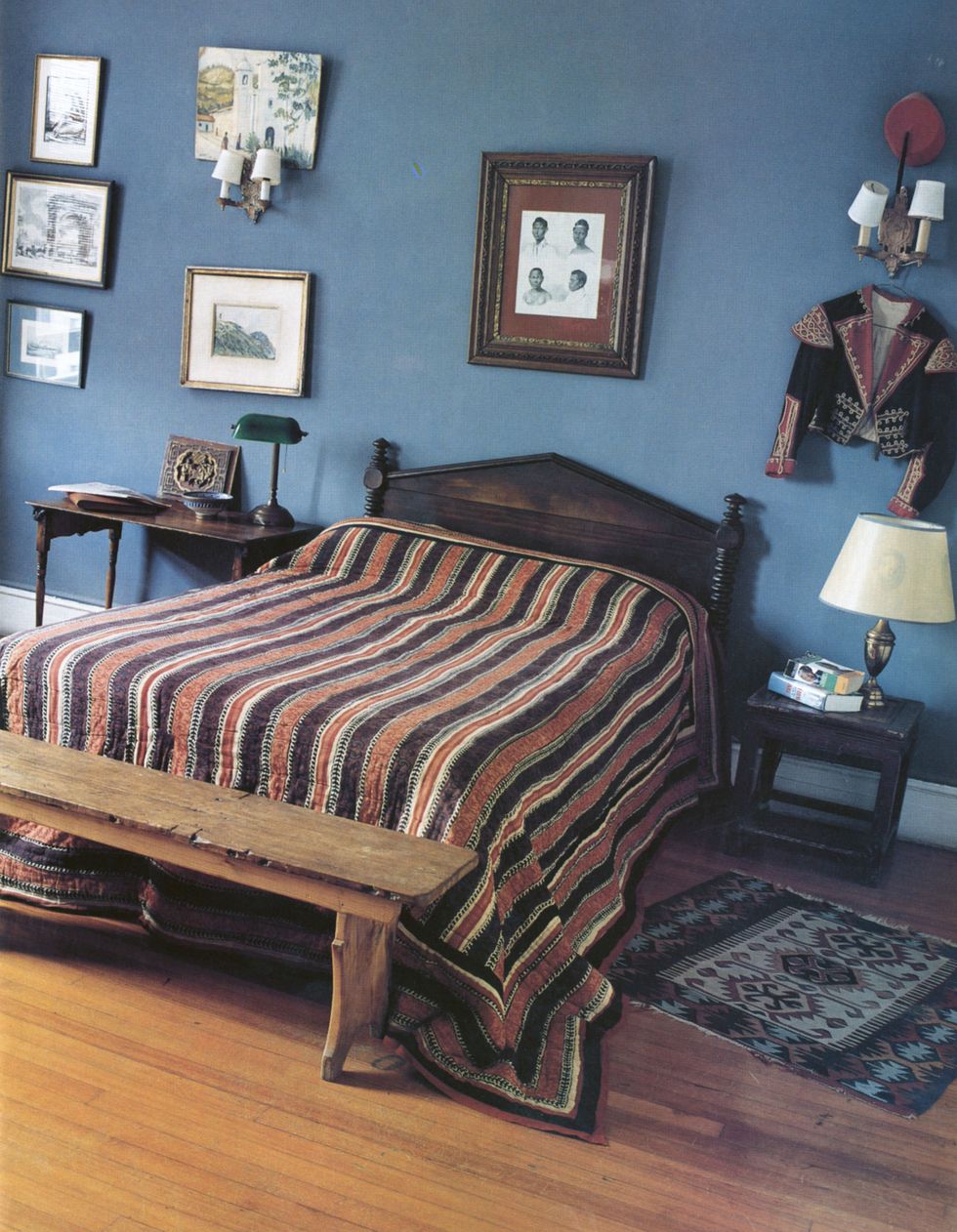 blue painted bedroom with objects hanging from the wall including a studded embroidered matador jacket and some frames and a rustic bench and striped bedcover