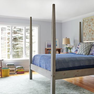 four poster bed in lightly dyed wood with a striped coverlet and multiple pills and two prints over the headboard and a rush seat chair across in the corner near a pile of books