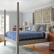 four poster bed in lightly dyed wood with a striped coverlet and multiple pills and two prints over the headboard and a rush seat chair across in the corner near a pile of books